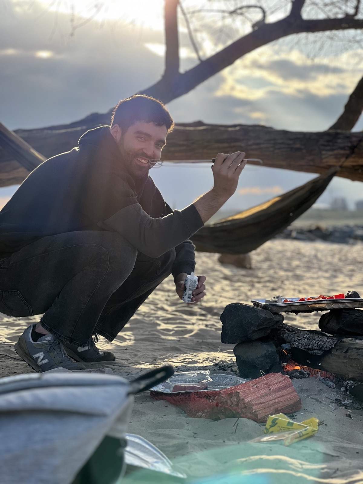 A cook tending a small fire on a beach at sunset, an old tree branch overhead.